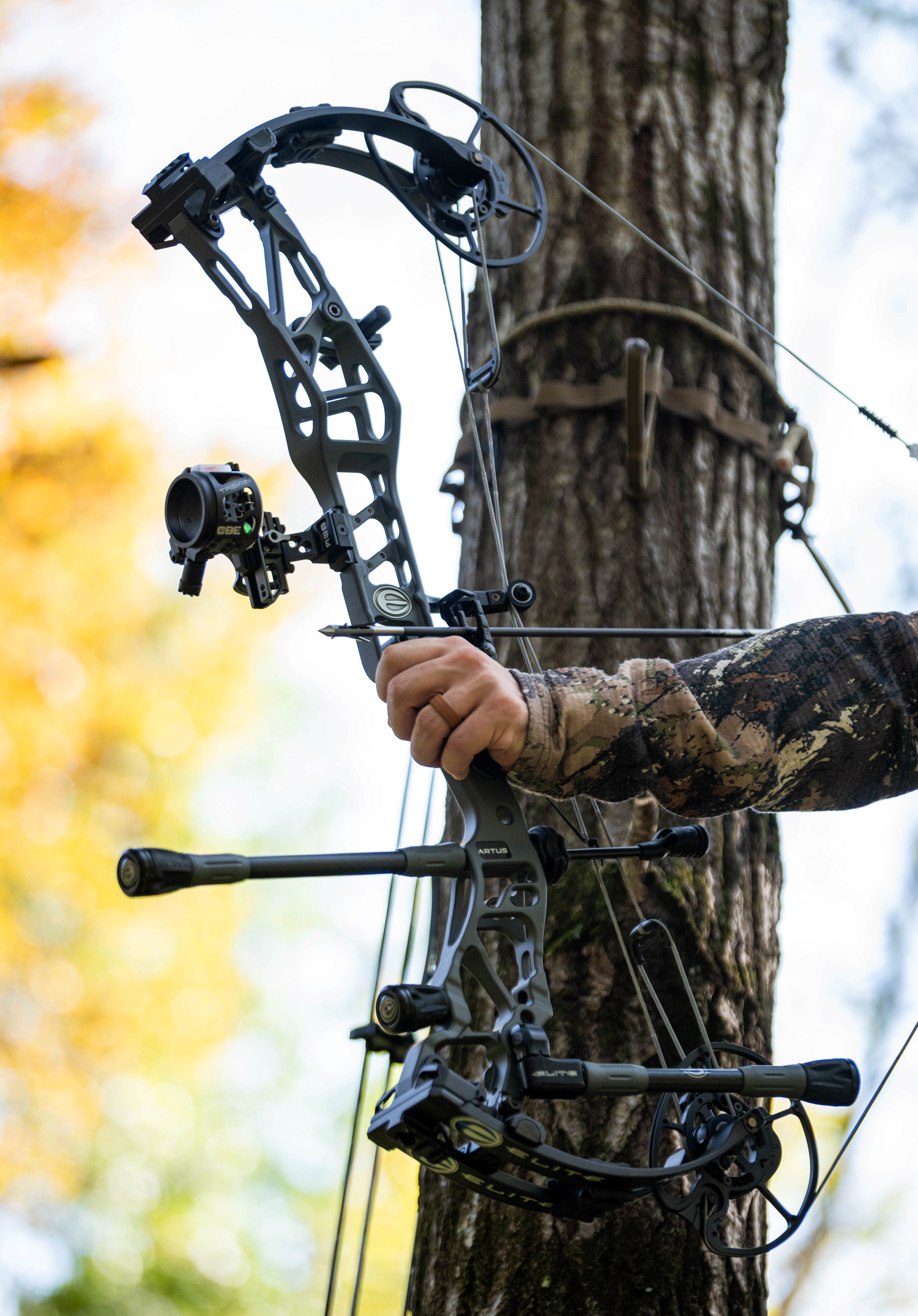 Archer drawing back an Elite Artus compound bow with a blurred tree background